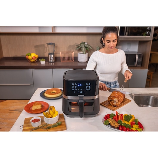 AIRFYER UNDER THE TABLE WITH: DESERT, POTATOS, CHICKEN, VEGETABLES AROUND. WOMAN HOLDING KNIFE TO SERVE CHICKEN.