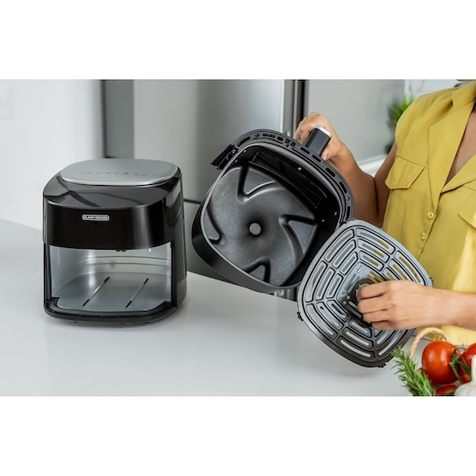 Digital AirFryer with front display and 360° technology. Airfryer under the table. Woman with a yellow T-shirt is checking the basket and the airfryer grill. the objective is to show the size inside the basket.