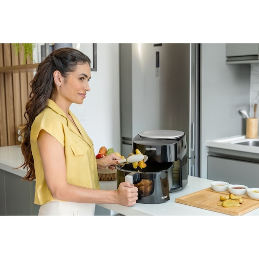 Digital AirFryer with front display and 360° technology. In a kitchen, a woman removes potatoes from the air fryer basket. The woman holds the potatoes with a tong in her left hand. The right hand is opening the basket. On the table there is a board with potatoes and sauces.