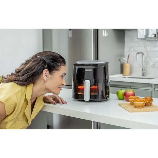 Digital AirFryer with front display and 360° technology. In a kitchen, a woman leans down to see the preparation of her cup cake inside the airfryer basket. Through the light inside the basket, it is possible to see the cup cake being prepared. The airfryer is on a table and next to the airfryer, two apples, one red and one green. And three cup cakes ready under the cutting board.
