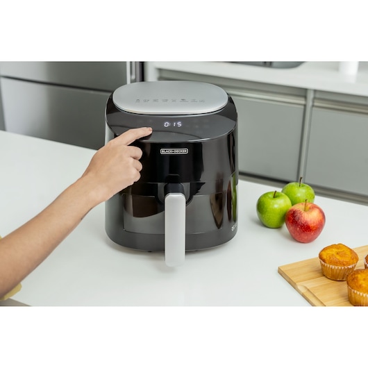 Digital AirFryer with front display and 360° technology. In a Kitchen, woman selects the airfryer's digital panel with her finger. The airfryer is on a table and next to it, 3 apples, 2 green, 1 red. Also next to it are 2 cup cakes on a cutting board.