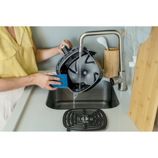 Digital AirFryer with front display and 360° technology. In a kitchen, a woman washes the basket and the airfryer rack. The tap is open and the blue washing sponge is next to the basket, simulating washing the product.