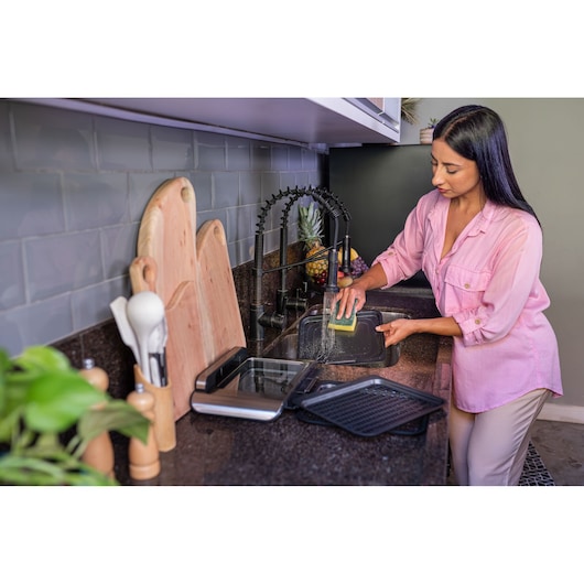 Woman, black hair, pink shirt, washing the oven tray in the sink, to show the ease of washing and non-stick