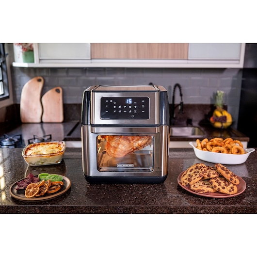 Airfryer in the center of the counter with chicken being prepared. It is possible to check the inside of the oven thanks to the internal lighting and glass display. On the side, ready: cookies, fresh fruit, roasted onions and cauliflower in white sauce