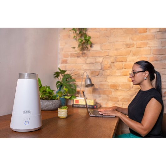 While a woman works in her home office, the air humidifier is being used, with snow coming out of the product