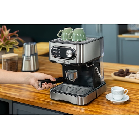 Espresso maker in a kitchen. Woman holds a measuring spoon and leveler, positioning it close to the machine. Two green cups on top of the coffee maker, a white cup next to it.
