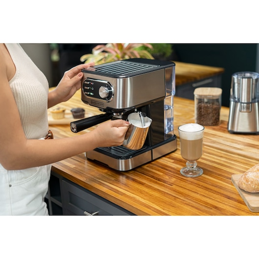 Woman holds glass to froth milk in coffee machine. Next to it is a ready-made cappuccino.