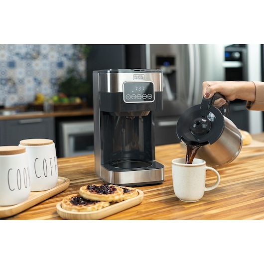Programmable electric coffee maker under a wooden countertop. Woman removes stainless steel jug from the coffee maker and pours coffee into a mug. Next 3 waffles with jam