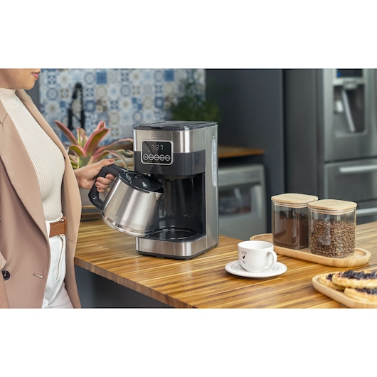 Woman places the stainless steel on the base of the programmable coffee maker, with a cup of powder and coffee beans next to it.
