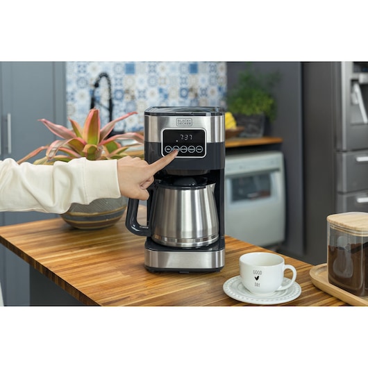 Woman selects the programming option on the coffee maker's digital panel. The coffee maker is positioned on the wooden table, next to a cup, coffee and a plant behind.