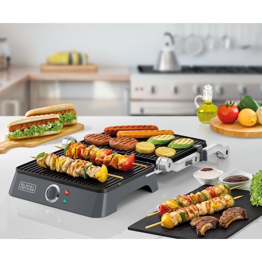 grill G1400 BLACK and Decker. Open 180º  under the table with many options of food. There are food under the product as barbecue, sausage, vegetables. Under the table are hot dogs, barbecue salse and 2 skewer