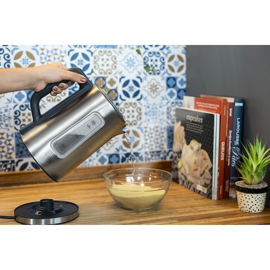 Programmable Electric Kettle. On a wooden countertop in the kitchen, a woman uses the kettle to put water in a bowl with food