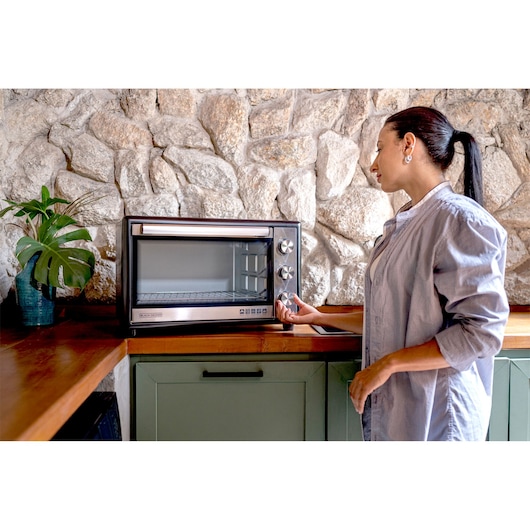 Photo of BLACKDECKER FT46 countertop oven on a wooden kitchen counter, person adjusting front knob, stone wall background.
