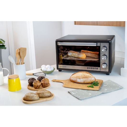 A BLACK+DECKER FT60 toaster oven on a kitchen counter, with bread inside baking. Surrounding the oven are various baked goods including muffins, bread rolls, and a loaf of bread on a wooden cutting board, as well as kitchen utensils, eggs, and a yellow jar.
