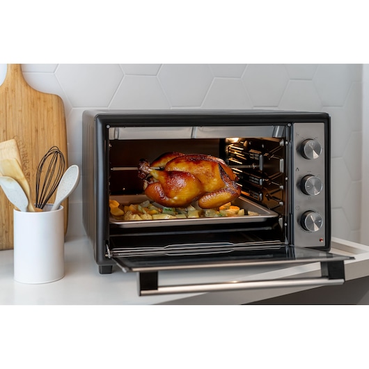 A BLACK+DECKER countertop oven in a kitchen, with a roasted chicken on a rotisserie inside and vegetables on a tray. Kitchen utensils and a wooden board are visible nearby.