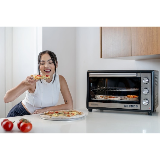 A BLACK+DECKER FT60 countertop oven is seen on a kitchen counter, with two pizzas inside it. In front of the oven, a person is eating a slice of pizza from a plate of pizza, with two tomatoes also visible on the counter. The person's face is blurred.