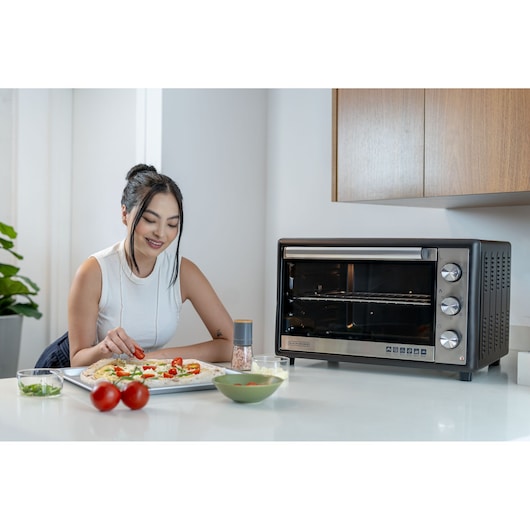 A person preparing food next to a BLACK+DECKER FT60 countertop oven in a modern kitchen. Fresh ingredients and kitchen utensils are on the counter.