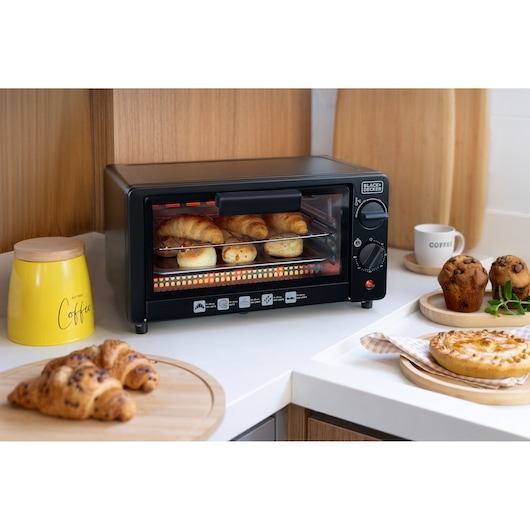 A BLACK+DECKER toaster oven on a kitchen counter, baking croissants and bread rolls. Surrounding the oven are baked goods, a yellow coffee canister, a cup labeled 'coffee', and wooden cutting boards.