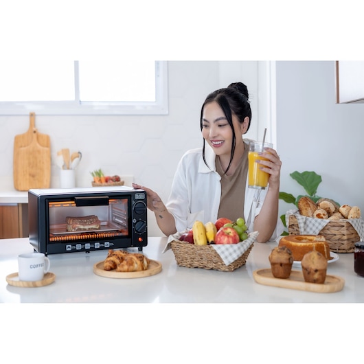 A BLACK+DECKER toaster oven is placed on a kitchen counter, with a sausage cooking inside. In front, there are plates and baskets with breakfast food including croissants, muffins, a loaf of bread, fruits like bananas, apples, and grapes, as well as a cup of coffee and a glass of orange juice held by a person. The kitchen background is bright and modern.