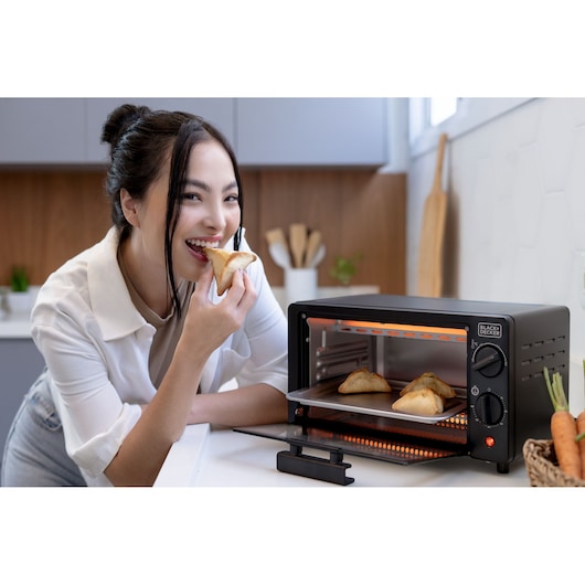 A person is eating a pastry in a kitchen while using a BLACK+DECKER toaster oven (model FT9N). The oven's door is open and inside there are baked pastries on a tray.