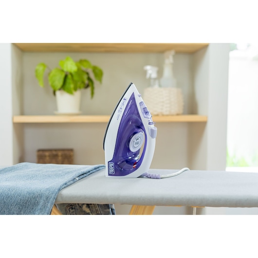 A BLACK+DECKER electric steam iron in purple and white, placed upright on an ironing board with denim fabric. The background includes shelves with a potted plant and household items.