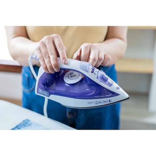 Photo of person holding BLACK+DECKER FX1050 steam iron with white and purple body while ironing clothes.