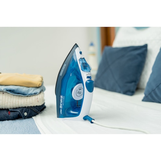 A BLACK+DECKER electric steam iron (model FX2150), blue and white, is placed upright on a bed beside neatly folded clothes and pillows in a bedroom setting.