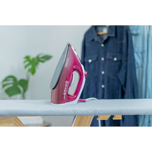 A close-up of a BLACK+DECKER electric iron in red color placed upright on an ironing board. In the background, there is a hanging denim shirt and a leafy green plant.