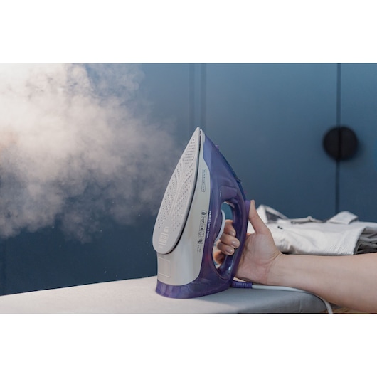 A hand holding a BLACK+DECKER steam iron, emitting steam, with clothes in the background on an ironing board.