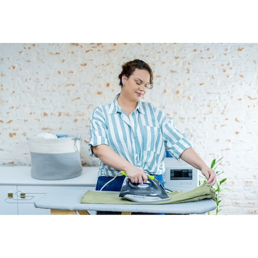 BLACKDECKER FX2750 steam iron in use on green pants, person ironing at home with basket and ironing board.