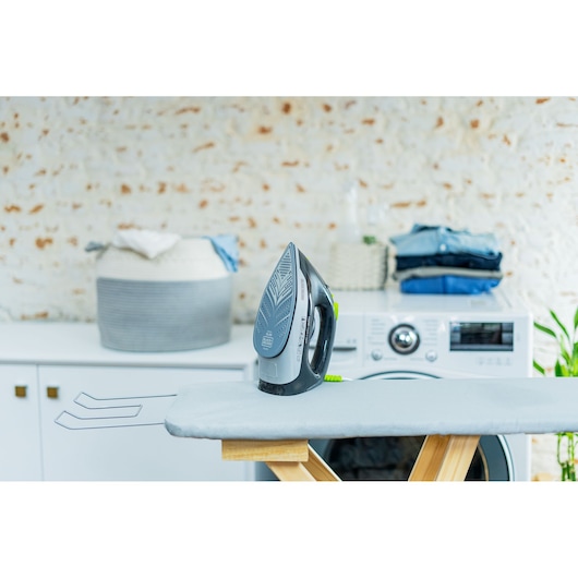 A BLACK+DECKER steam iron standing upright on an ironing board in a laundry room. In the background, there is a washing machine, a stack of folded clothes, and a laundry basket.