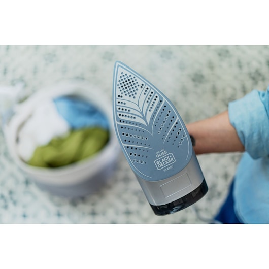 BLACK+DECKER FX2750 steam iron with ceramic soleplate, close-up view, person holding iron, laundry basket in background.