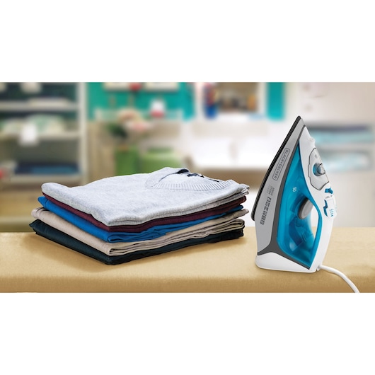 A blue and white BLACK+DECKER steam iron is placed upright on an ironing board next to a neatly folded stack of clothes in various colors. The background is a blurred laundry or household room.