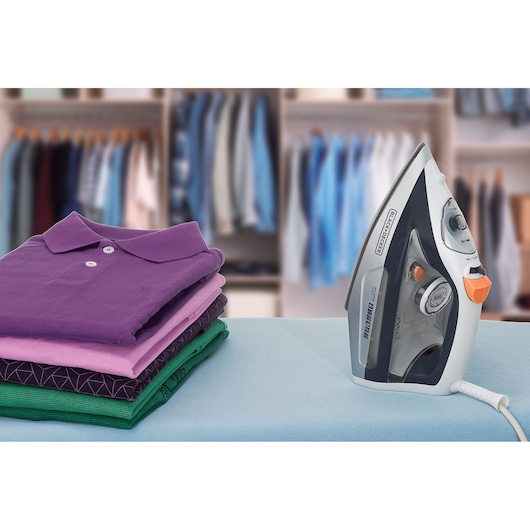 A BLACK+DECKER electric steam iron next to a stack of neatly folded shirts in various colors, placed on an ironing board with a closet in the background.