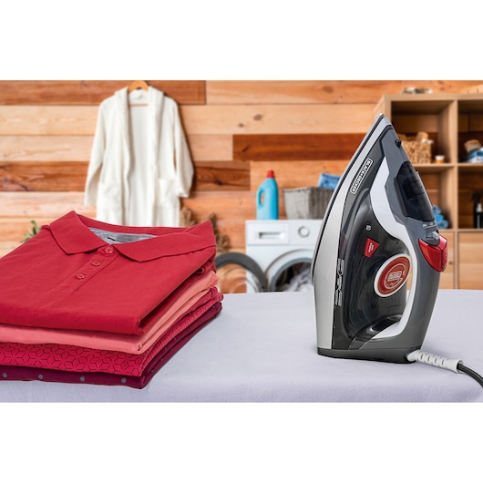 A BLACK+DECKER FX900 steam iron placed on an ironing board next to a stack of neatly folded shirts in a laundry room. The background shows wooden walls, shelves with laundry supplies, and a washing machine.