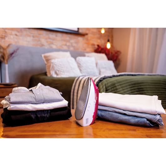 A BLACK+DECKER iron (model FXVG1) placed on a wooden table between two stacks of folded clothes in a cozy bedroom setting with a neatly made bed and decorative pillows in the background.