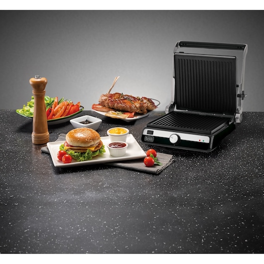 A BLACK+DECKER electric contact grill is shown open on a kitchen countertop, with food arranged around it including a hamburger with sauces, skewered meat, a salad, a pepper grinder, and some cherry tomatoes.