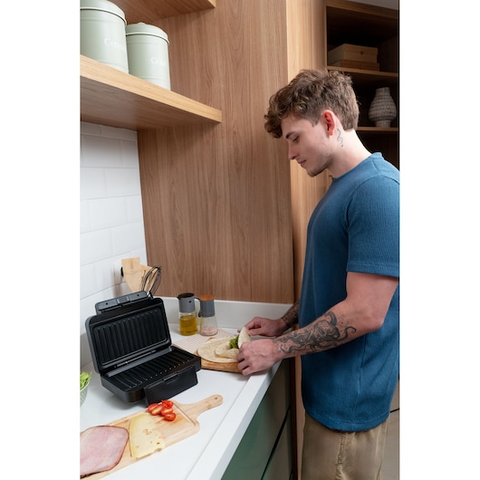 Person preparing a sandwich next to a BLACK+DECKER grill in a modern kitchen with wooden cabinets.