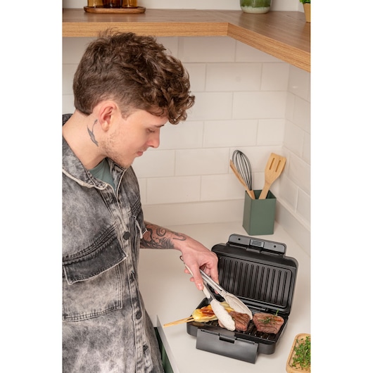 Person using a BLACKDECKER grill in a modern kitchen to cook meat and vegetables with tongs.