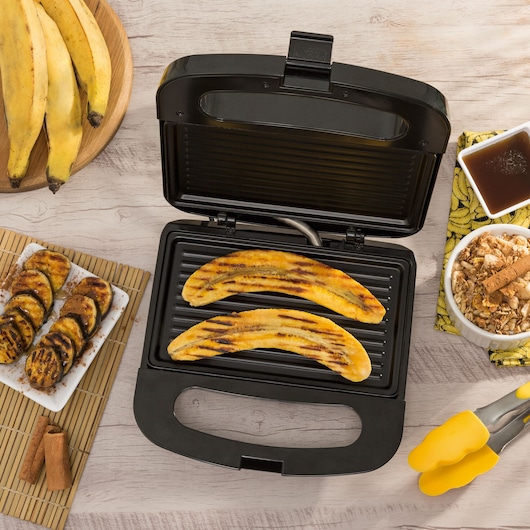 Two grilled plantains cooking in a Black+Decker indoor grill, surrounded by whole plantains on a wooden plate, grilled plantain slices on a white dish, a bowl of cereal with cinnamon sticks, a bowl of syrup, and yellow kitchen tongs on a light wooden table.