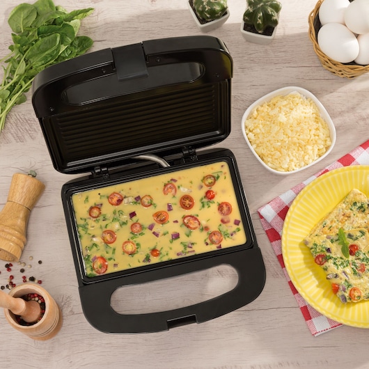A BLACK+DECKER electric grill with an open lid is filled with an egg and vegetable mixture, likely preparing an omelette. Around the grill, there are spinach leaves, a wooden pepper mill, a small bowl of shredded cheese, a basket of eggs, a yellow plate with a cooked omelette, and two small decorative cacti.