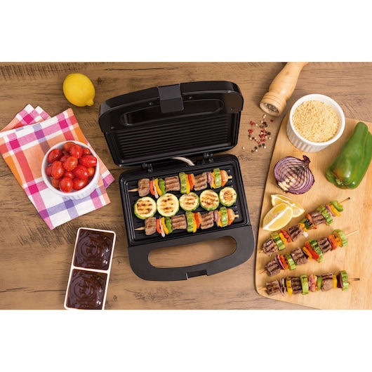 A BLACK+DECKER electric grill is open on a wooden table, grilling skewers with pieces of meat and vegetables. Next to the grill are ingredients such as cherry tomatoes, barbecue sauce, couscous, lemon wedges, green bell pepper, red onion, and skewers arranged on a cutting board.