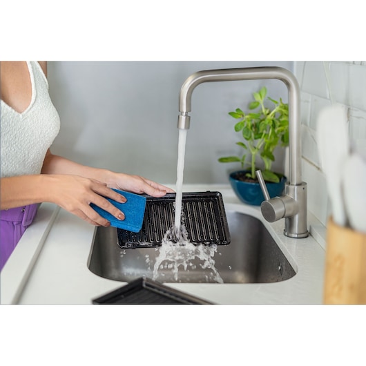 BLACKDECKER GW750 grill plate being washed in a kitchen sink with running water and blue sponge.