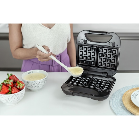 BLACKDECKER GW750 waffle maker in use on kitchen counter, person pouring batter into open plates, strawberries nearby.
