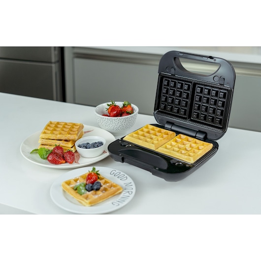 A BLACKDECKER waffle maker is shown on a kitchen counter, with two freshly cooked waffles inside. The scene also includes plates with waffles, strawberries, blueberries, and mint leaves, along with two small bowls of fruit.