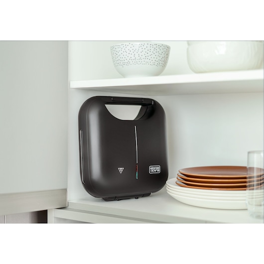 A BLACK+DECKER sandwich maker, model GW750, is placed vertically on a kitchen shelf next to stacked plates and bowls.
