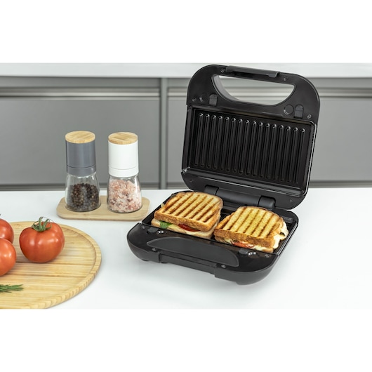 A BLACKDECKER sandwich maker with two grilled sandwiches inside, placed on a kitchen counter. Nearby are a wooden tray with fresh tomatoes and a sprig of rosemary, and a set of salt and pepper grinders.