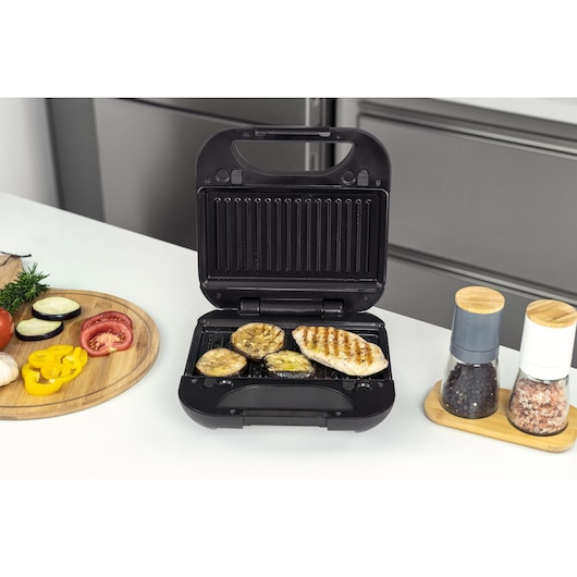 A black electric grill with a grilled chicken breast and vegetables on the cooking plate. Nearby are a round wooden board with sliced vegetables, and two spice grinders on a small tray. The grill is open and placed on a white countertop in a modern kitchen.