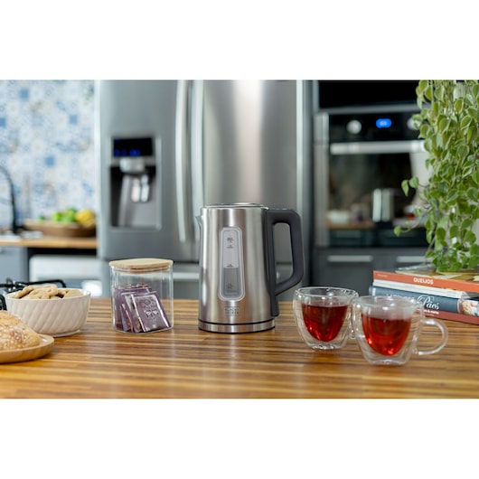 A Black+Decker electric kettle is placed on a wooden kitchen counter, surrounded by a bowl of cookies, a jar of tea bags, two clear glass cups filled with tea, and some books. The background shows a modern kitchen with a refrigerator, oven, and some greenery.