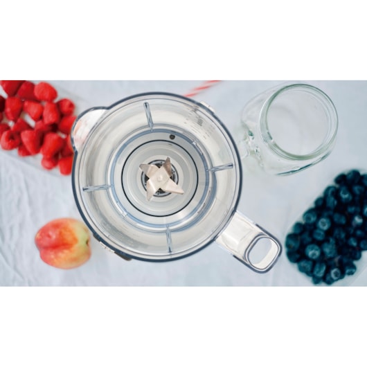 Top view of a blender jar with metal blades in the center, surrounded by ingredients including strawberries, blueberries, and an apple, along with a clear glass cup on a white surface.